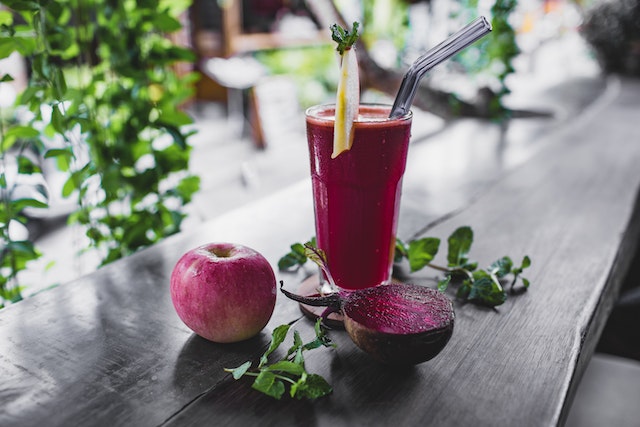 A glass filled with purple-looking liquid beside an apple and a sliced beetroot