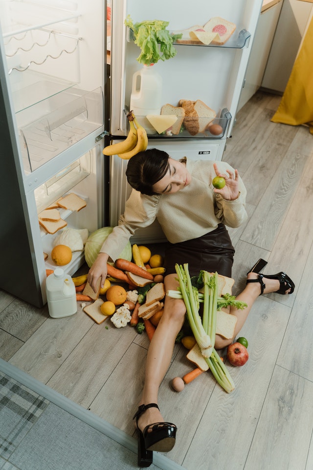 Woman sitting beside an open fridge with a bunch of vegetables and fruits around her.