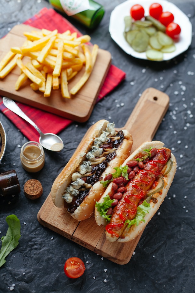 Hot dogs and buns served on a wooden board with fries and sliced fruits on the side