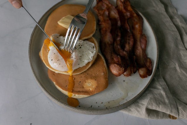 Hands slicing into a piece of poached egg on tops of pancakes with fried bacon on the side