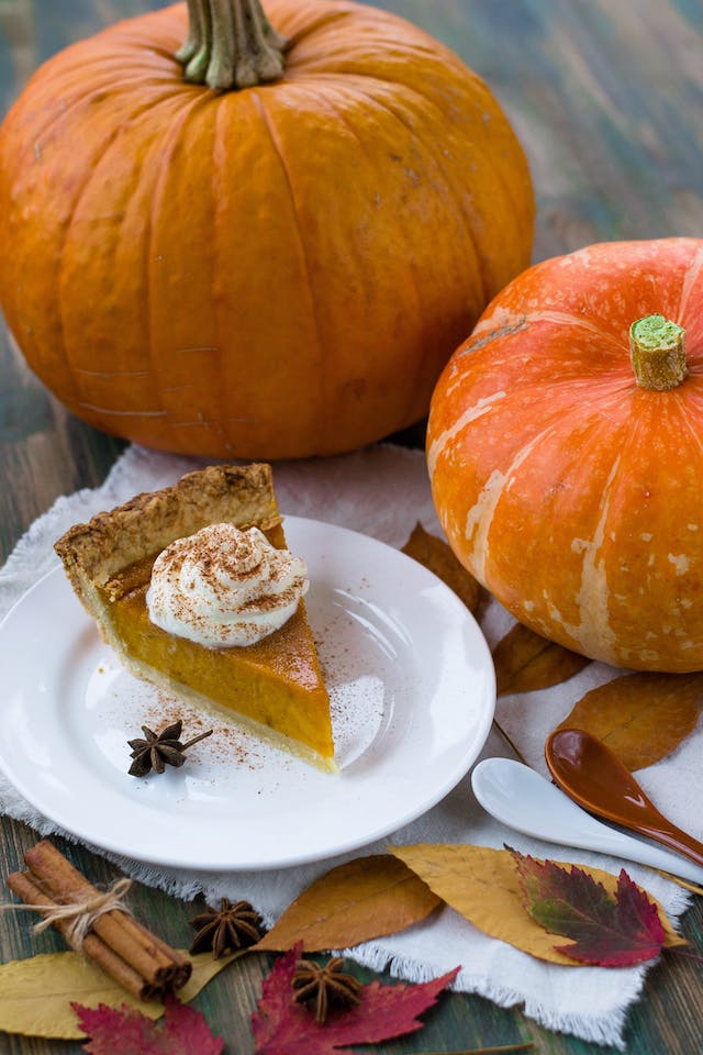 A slice of pumpkin pie on a white ceramic plate beside two pumpkins